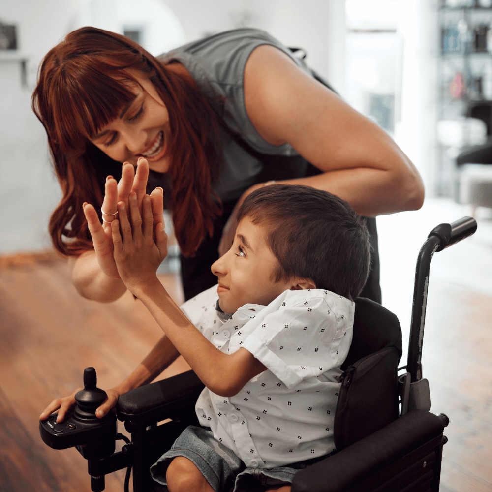 A smiling woman bends down to give a high five to a young boy in a wheelchair who is looking up at her, both appearing happy and engaged indoors.