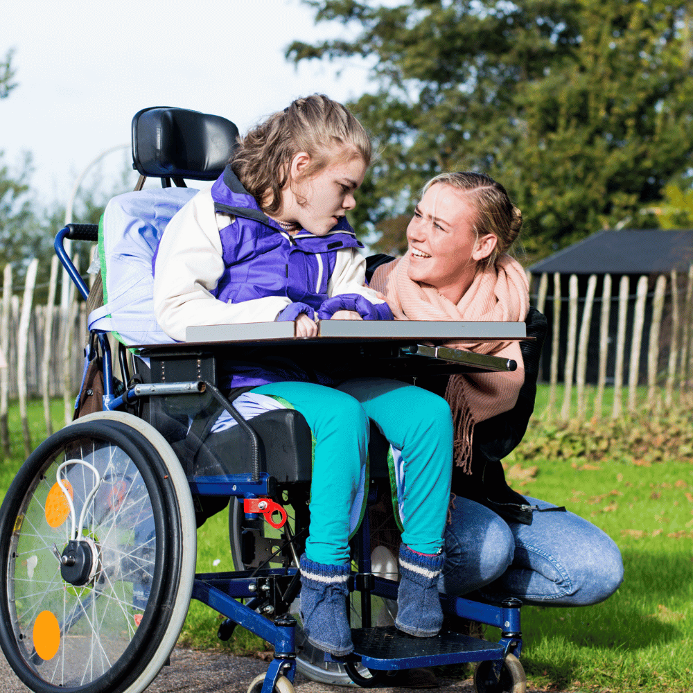 A young girl in a wheelchair sits outdoors with a woman kneeling beside her. They are both smiling and making eye contact, surrounded by greenery and a wooden fence in the background.