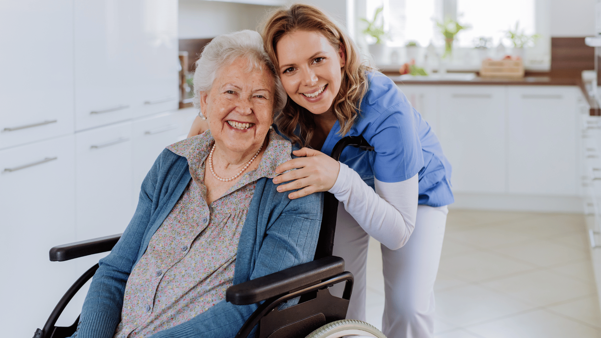 A smiling elderly woman in a wheelchair is embraced by a cheerful young caregiver in blue scrubs, both looking at the camera in a bright, modern kitchen.