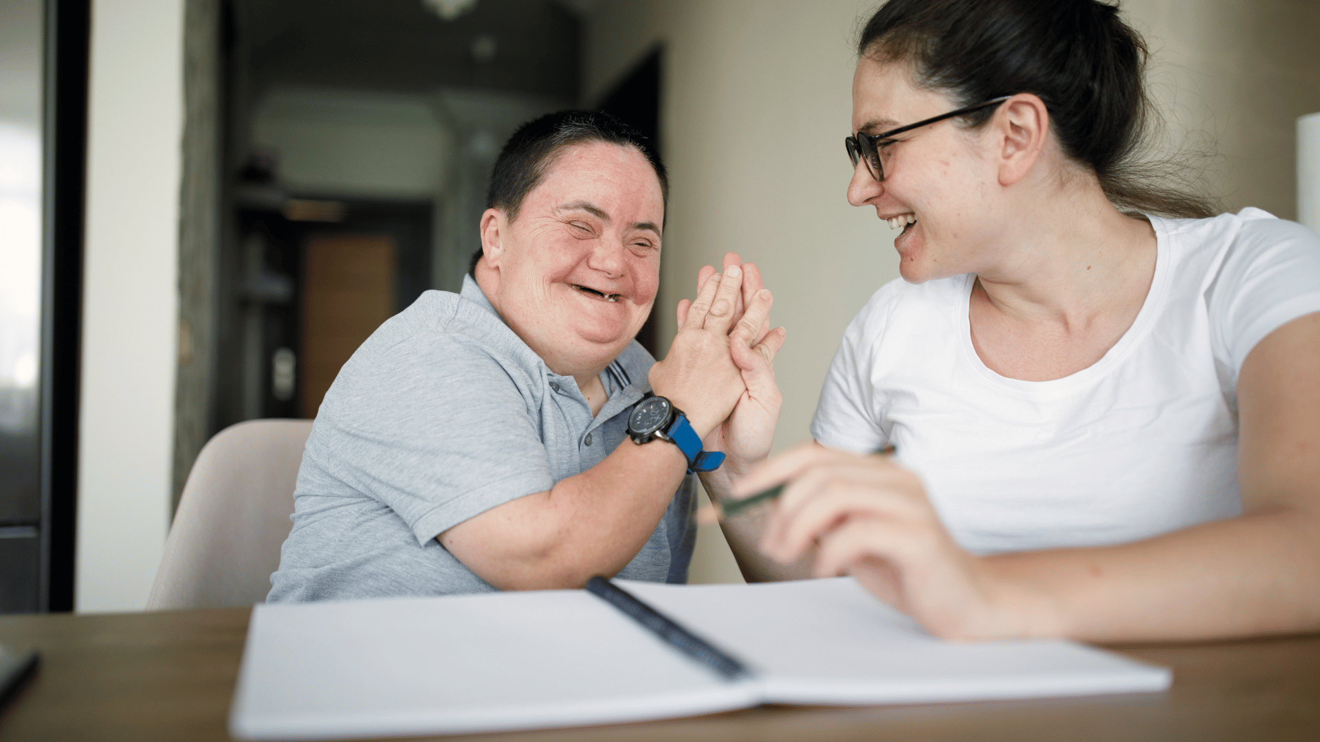 Two people sit at a table laughing and smiling together. One is a woman wearing glasses and a white shirt, holding a pen, while the other is a person with Down syndrome, wearing a gray shirt, clapping their hands happily.