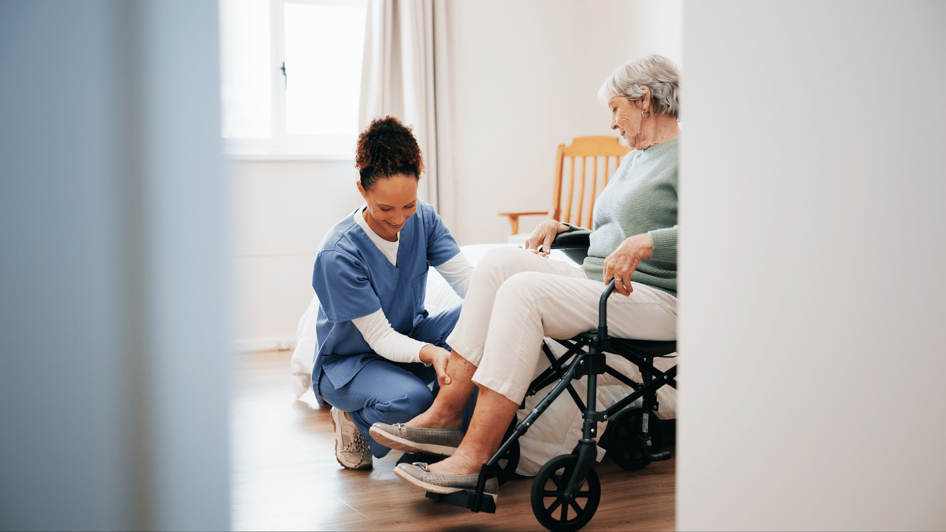 A nurse in blue scrubs assists an elderly woman in a wheelchair, helping her put on slippers in a bright, tidy room with a bed and wooden chair visible.