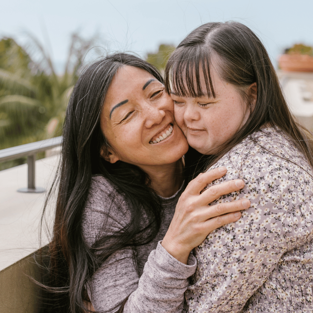 A woman with long dark hair lovingly hugs a young girl with Down syndrome. Both are smiling and wearing patterned sweaters, standing outdoors near a railing with trees in the background.