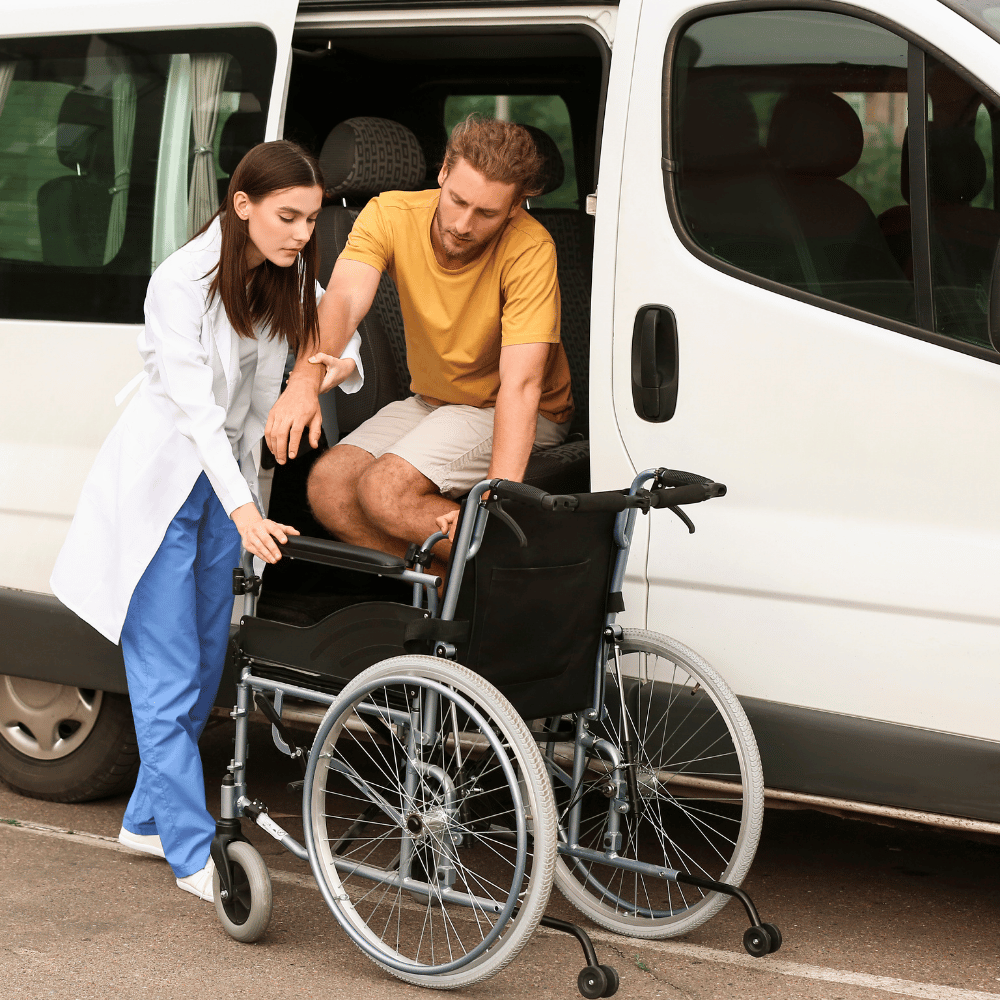 A woman in medical attire assists a man in a yellow shirt as he transfers from a van into a wheelchair parked beside the vehicle.